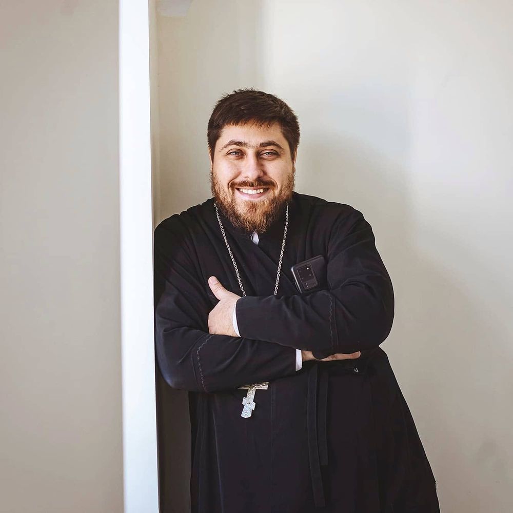 Священник Павел Сергеев улыбается, в подряснике с телефоном в руке